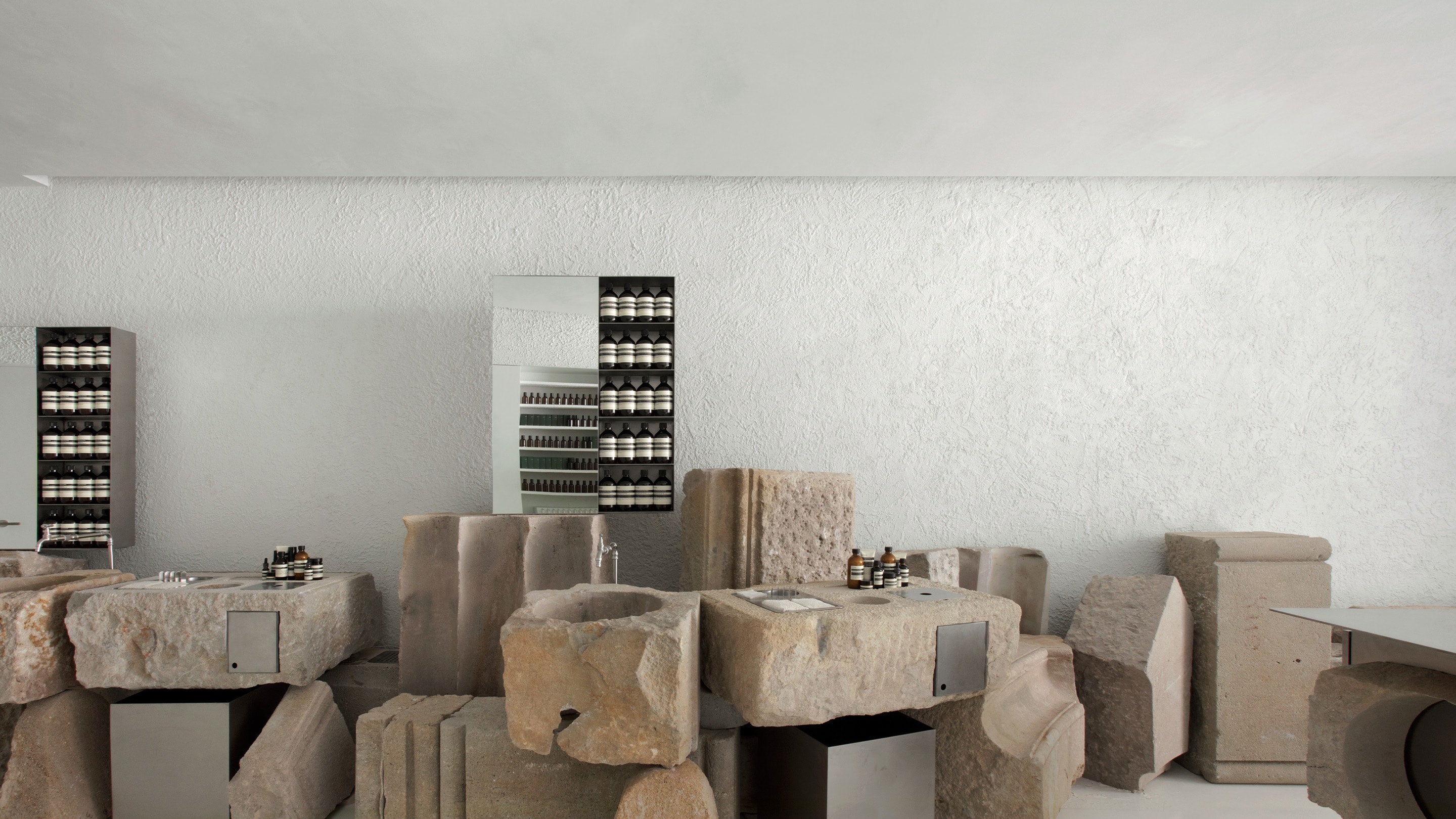 Aesop Diagonal store interior, carved stone basin and metal shelving displaying rows of amber bottles.