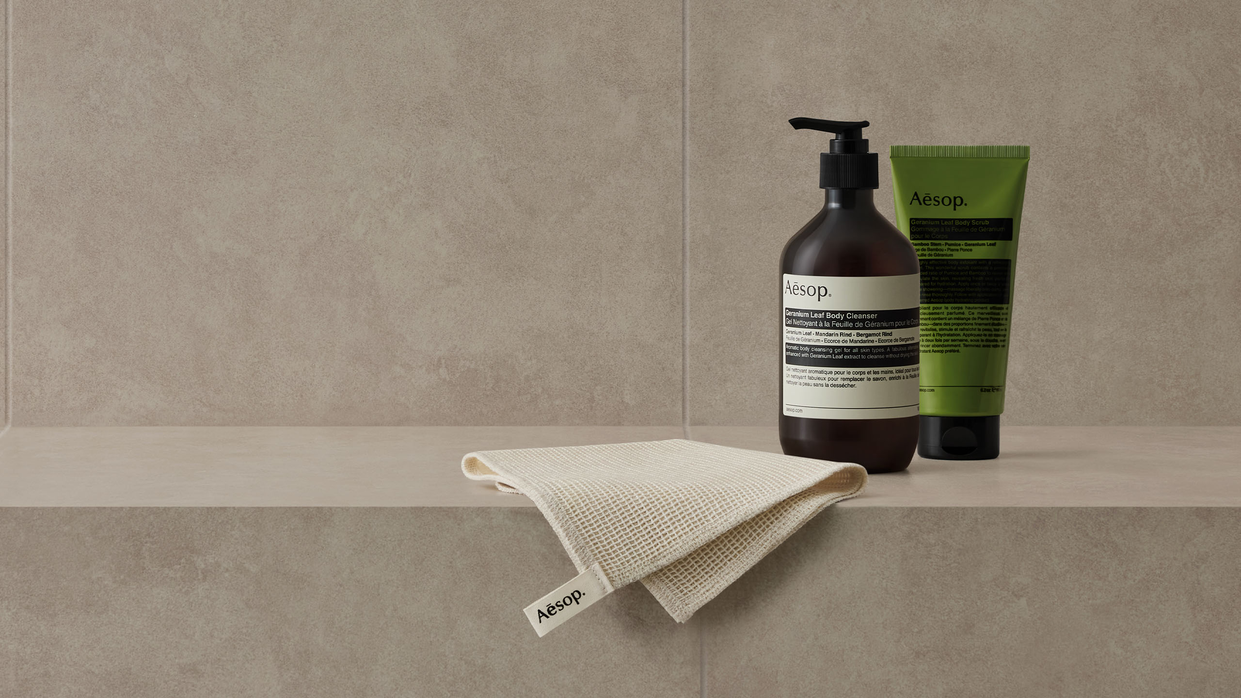 Geranium Leaf Body Cleanser and Geranium Leaf Body Scrub alongside Aesop shower towel placed on the grey bathroom tile. 
