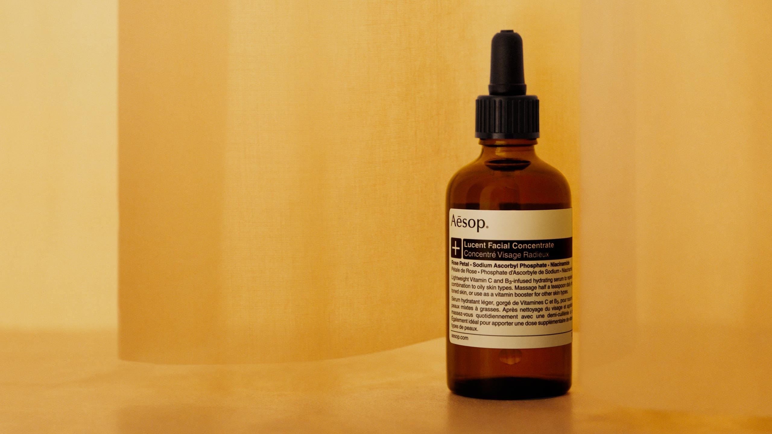 Aesop Lucent Facial Concentrate in amber glass bottle with pipette dropper placed in front of yellow-orange background