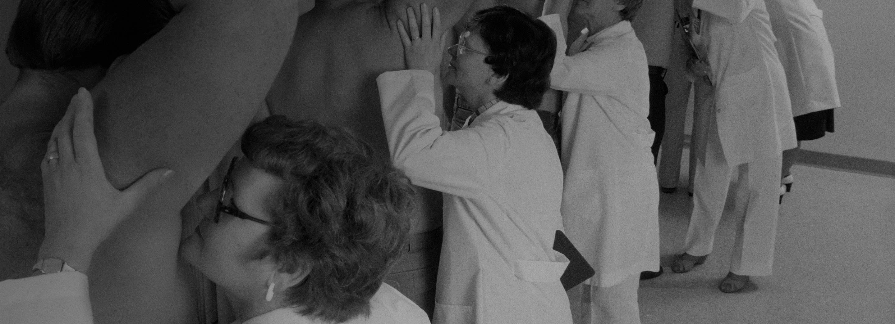 Retro black and white photography of women in lab coats smelling the armpits of a line-up of shirtless men.