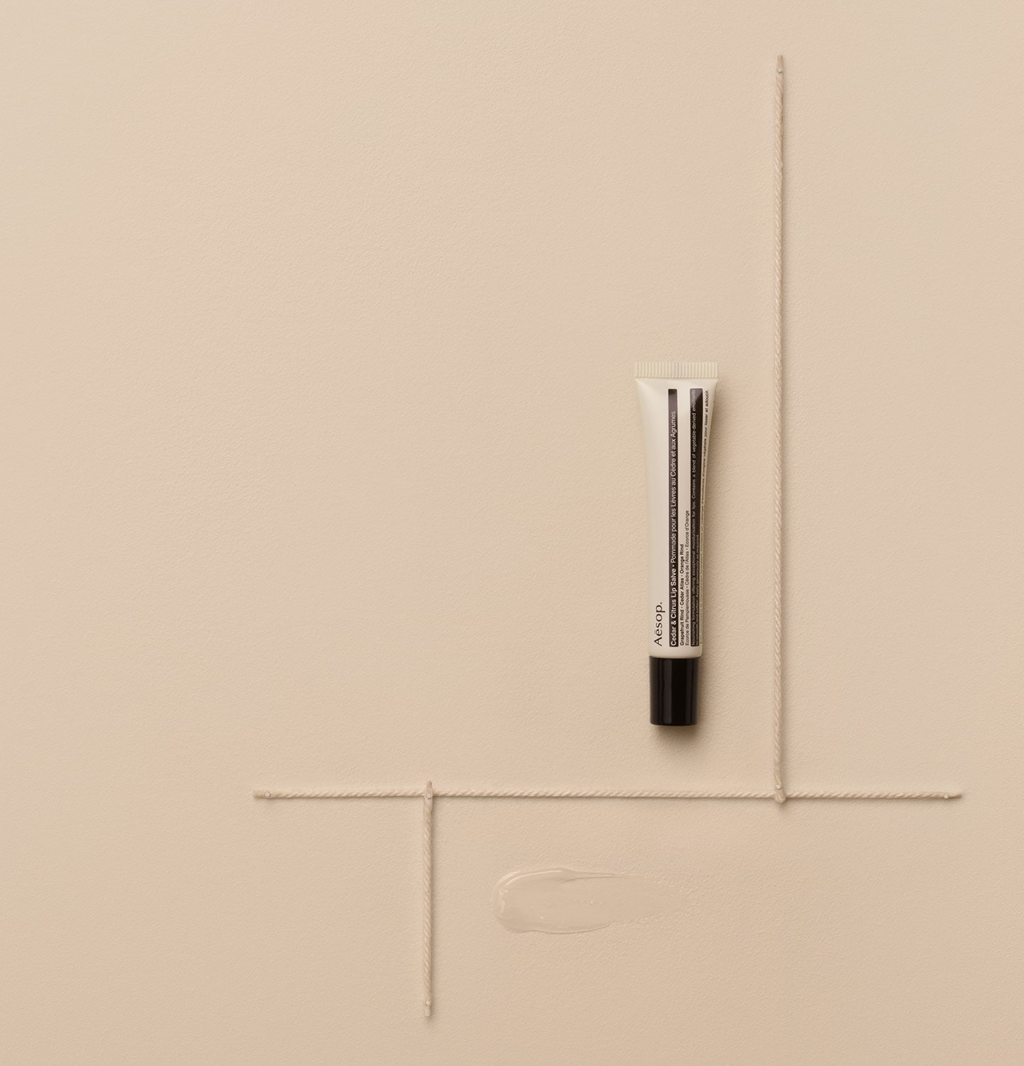 Aesop Cedar & Citrus Lip Salve in a tube arranged alongside cotton cords on a beige textured surface.