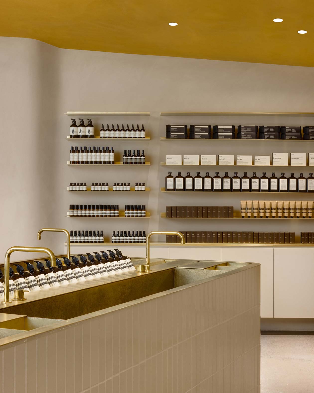A sink fitted with a brass tap and countertop in the foreground; shelves of Aesop product are neatly arranged on the shelves in the background.