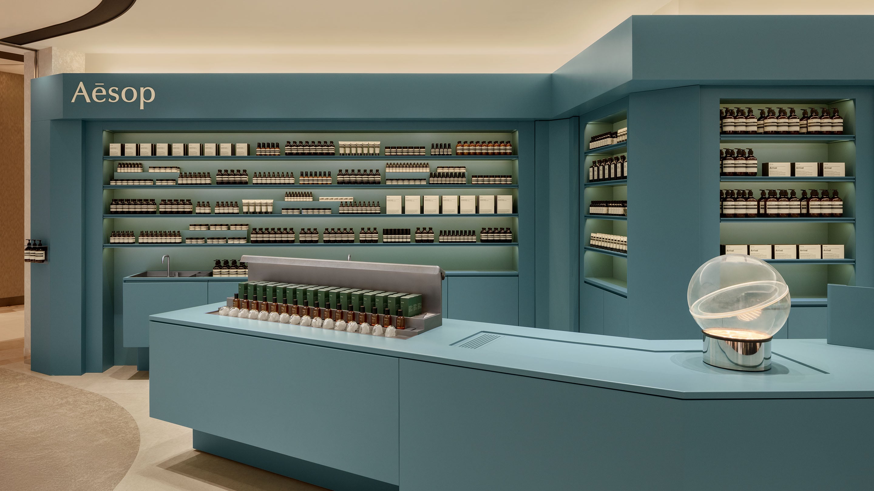 Fufuoka airport store interior featuring a turquoise color scheme, lined with shelves displaying Aesop products. A sleek counter is positioned in the foreground, with a decorative glass orb on it. 