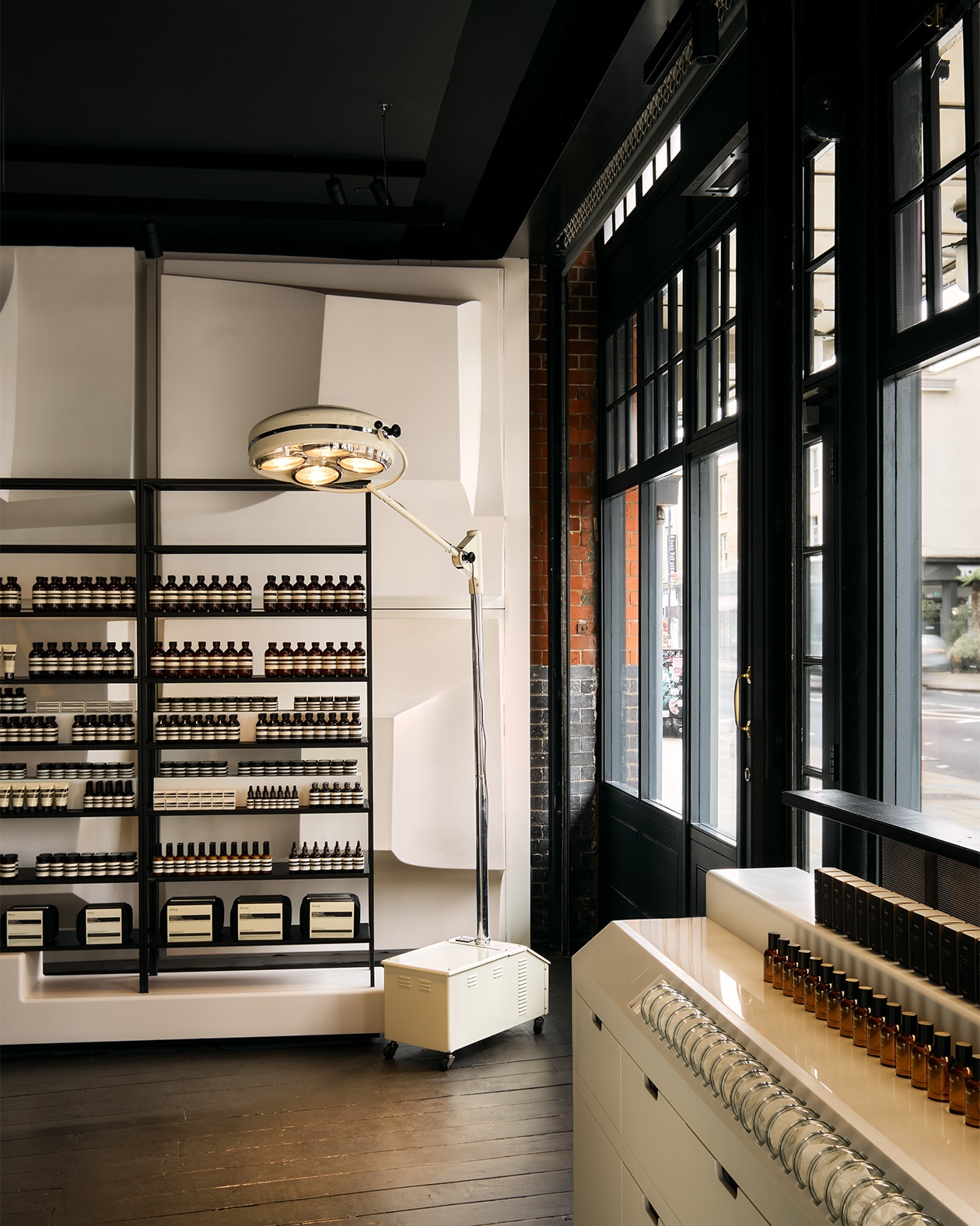 Aesop Spitalfields store interior entrance painted in black, contrasting with white feature walls. Lamp feature sits in the corner of the room and amber bottle product displays sit on custom shelving.